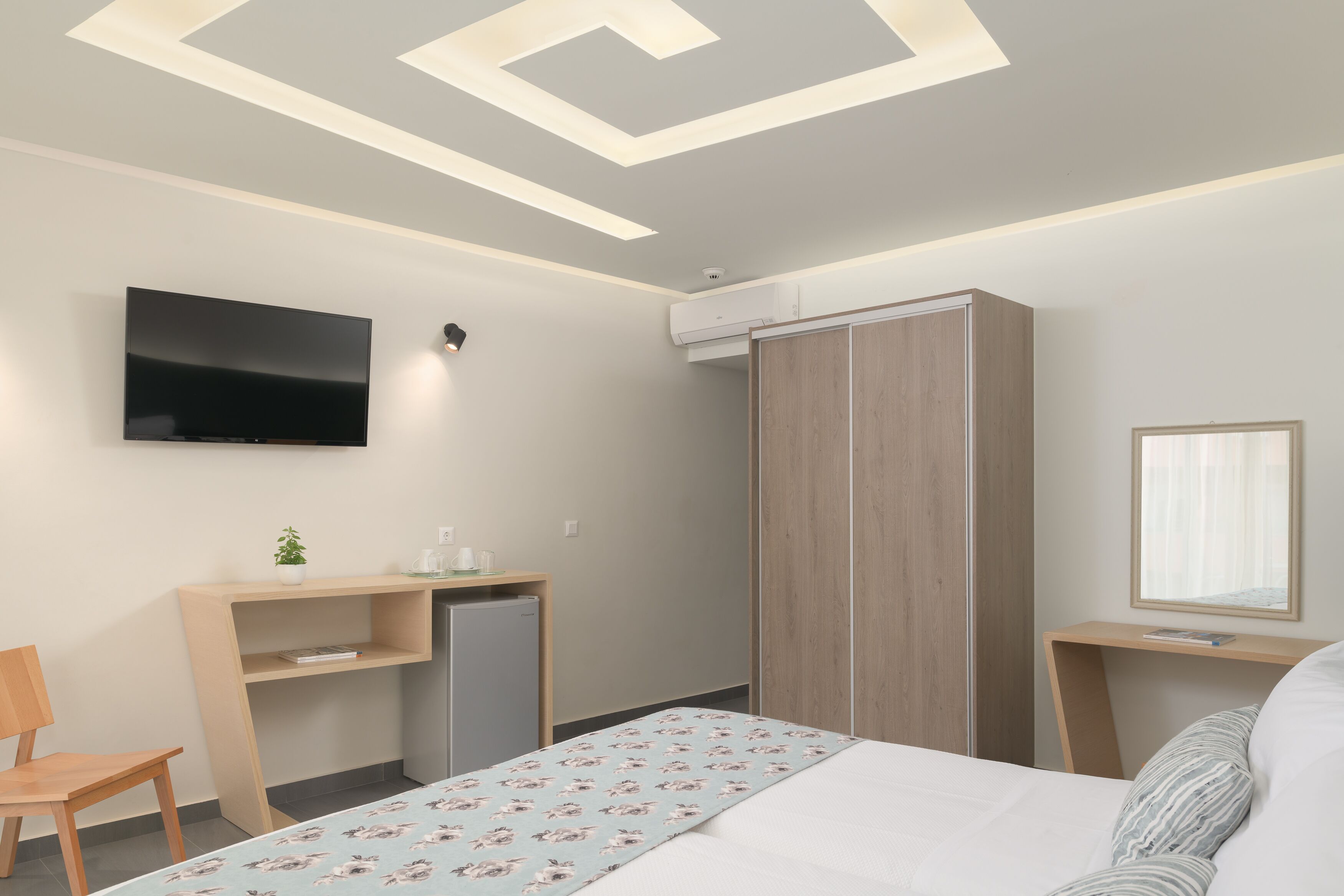 A modern hotel room with a bed, a desk with a computer, a TV mounted on the wall, and a wardrobe.