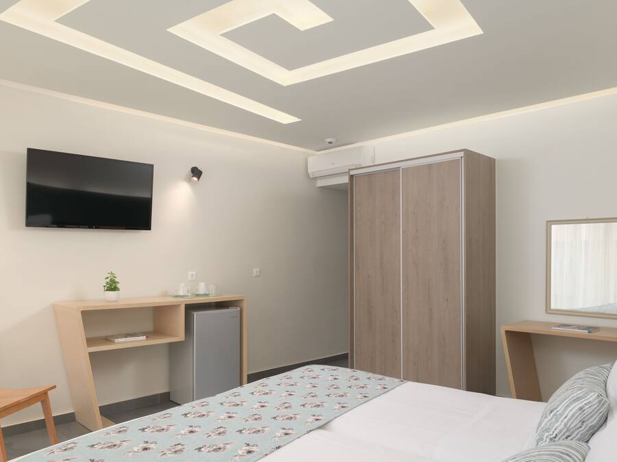 A modern hotel room with a bed, a desk with a computer, a TV mounted on the wall, and a wardrobe.
