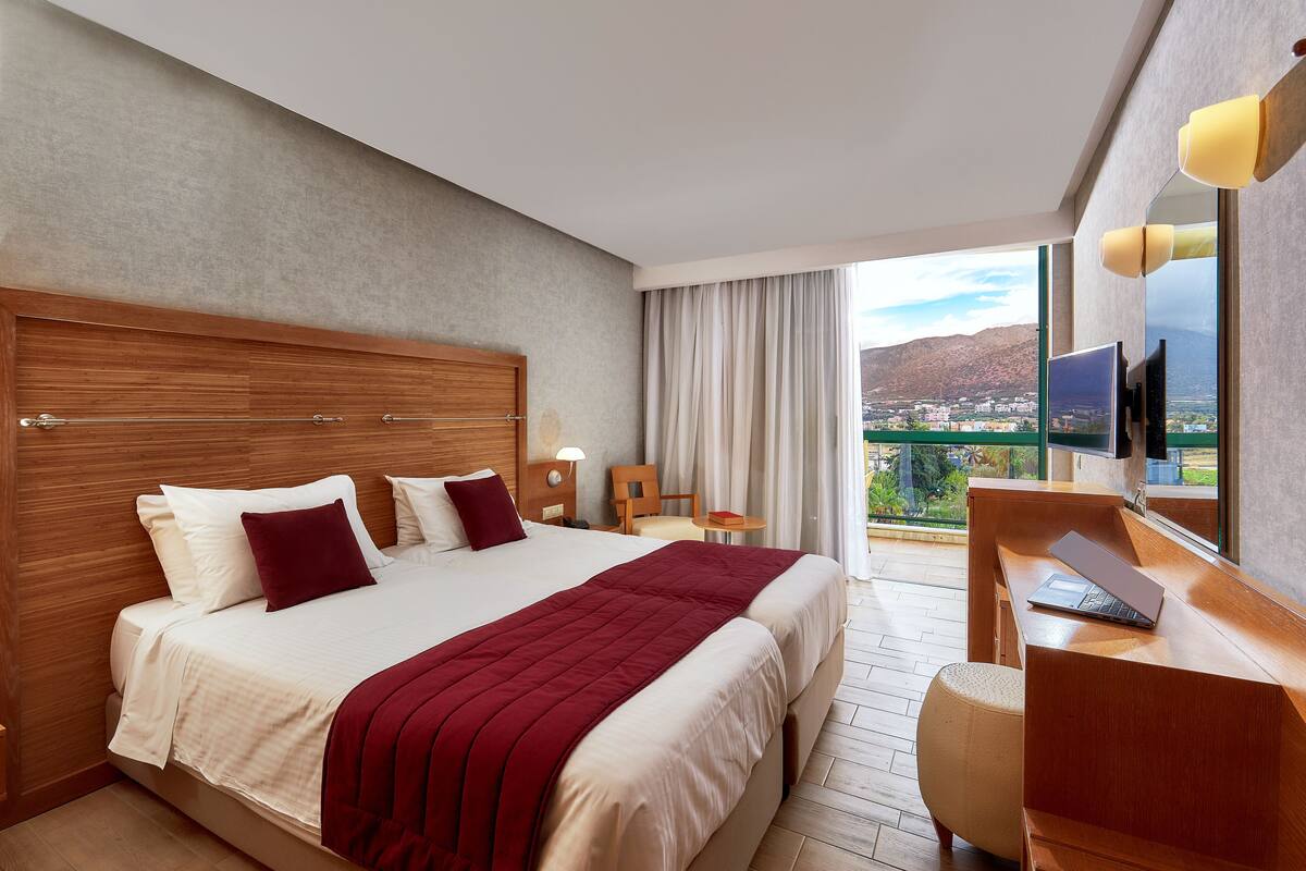 A hotel room with a large bed, a desk with a laptop, a chair, a TV, and a balcony with a city view.