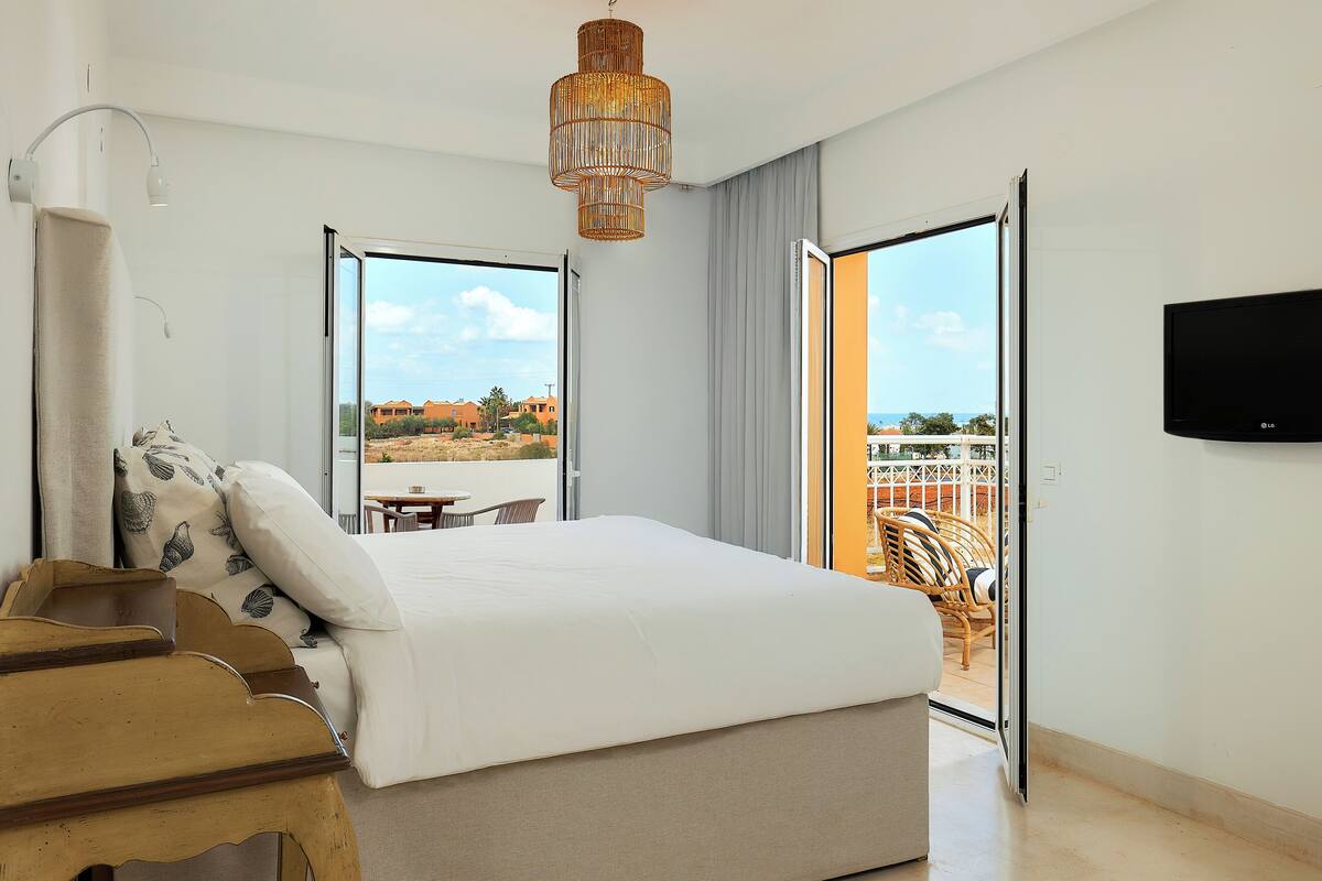 A hotel room with a bed, a wooden side table, a television, and a balcony with a view of buildings and a palm tree.