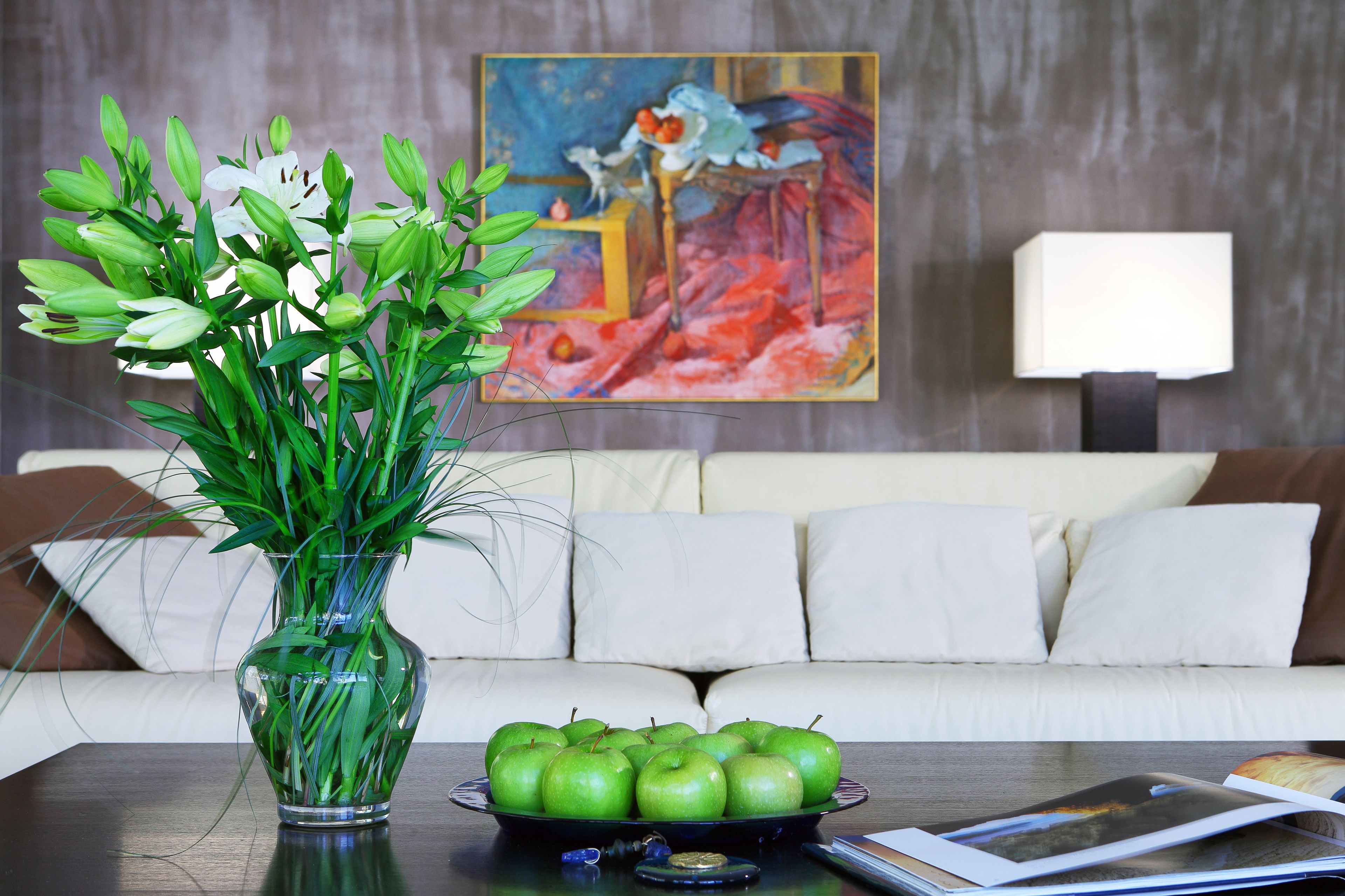A living room with a vase of green plants, a bowl of green apples, and a painting on the wall.