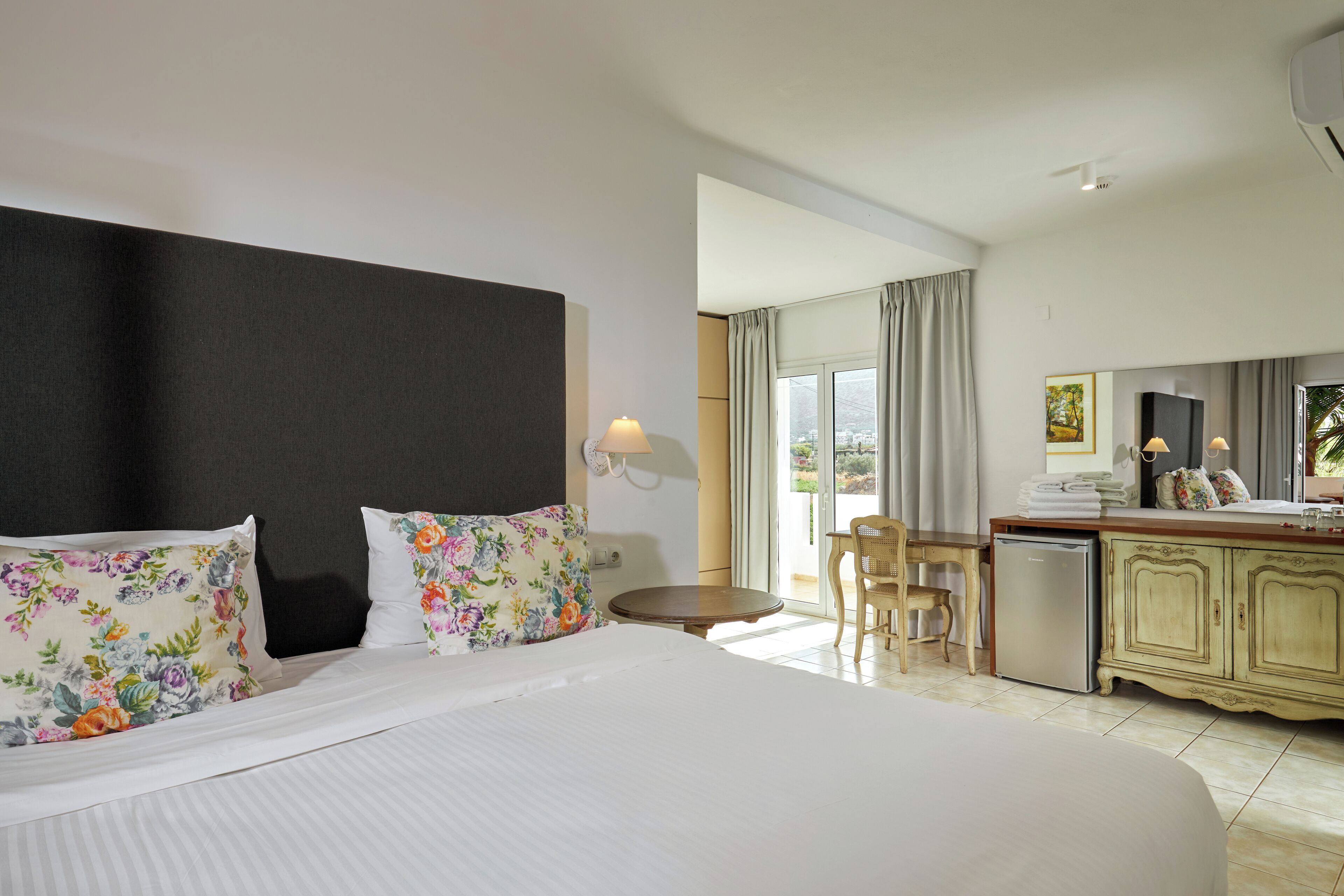 A hotel room with a large bed, a desk with a chair, a small refrigerator, and a view of the outdoors.