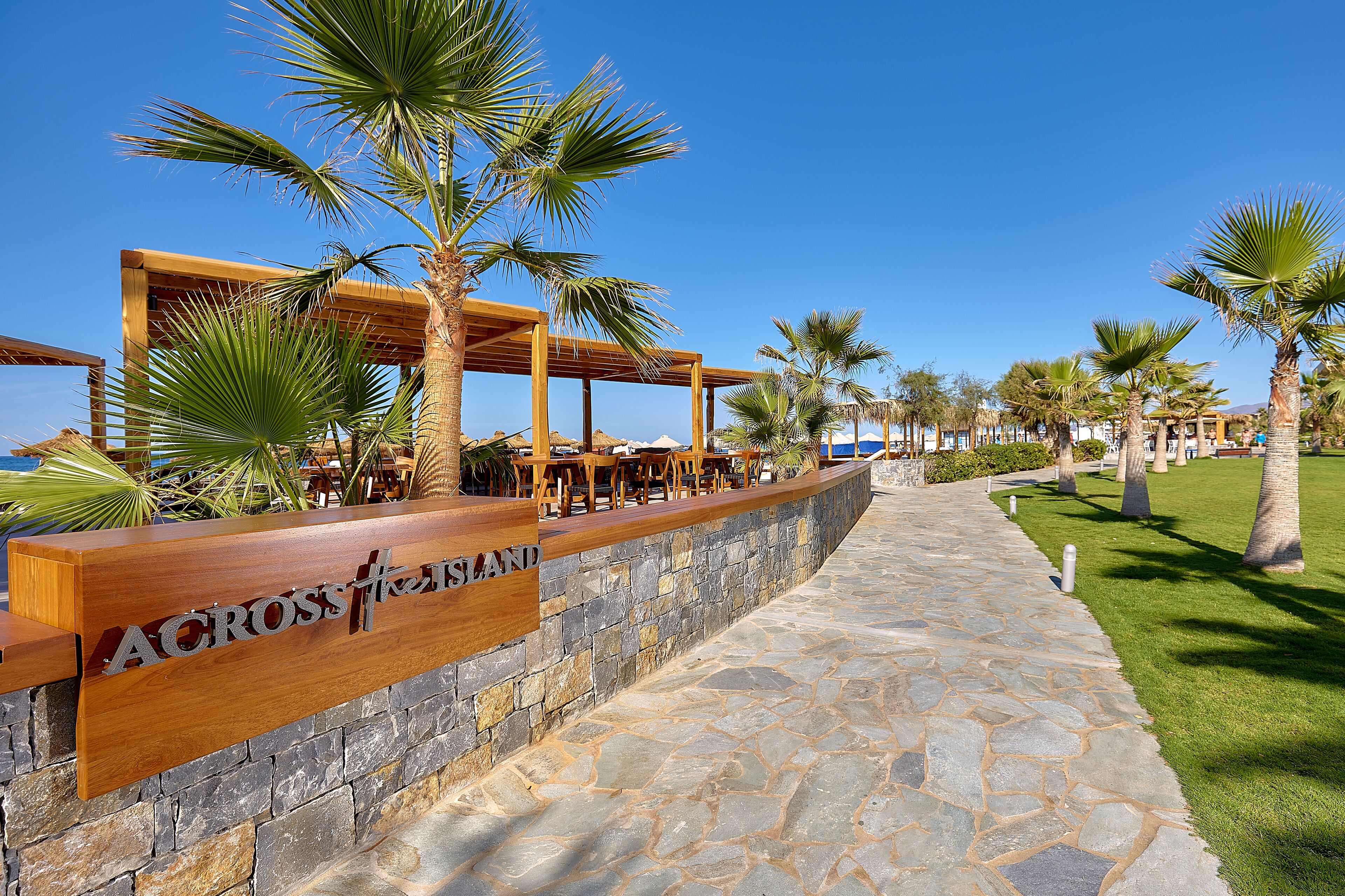 A wooden outdoor seating area with a stone wall and palm trees.