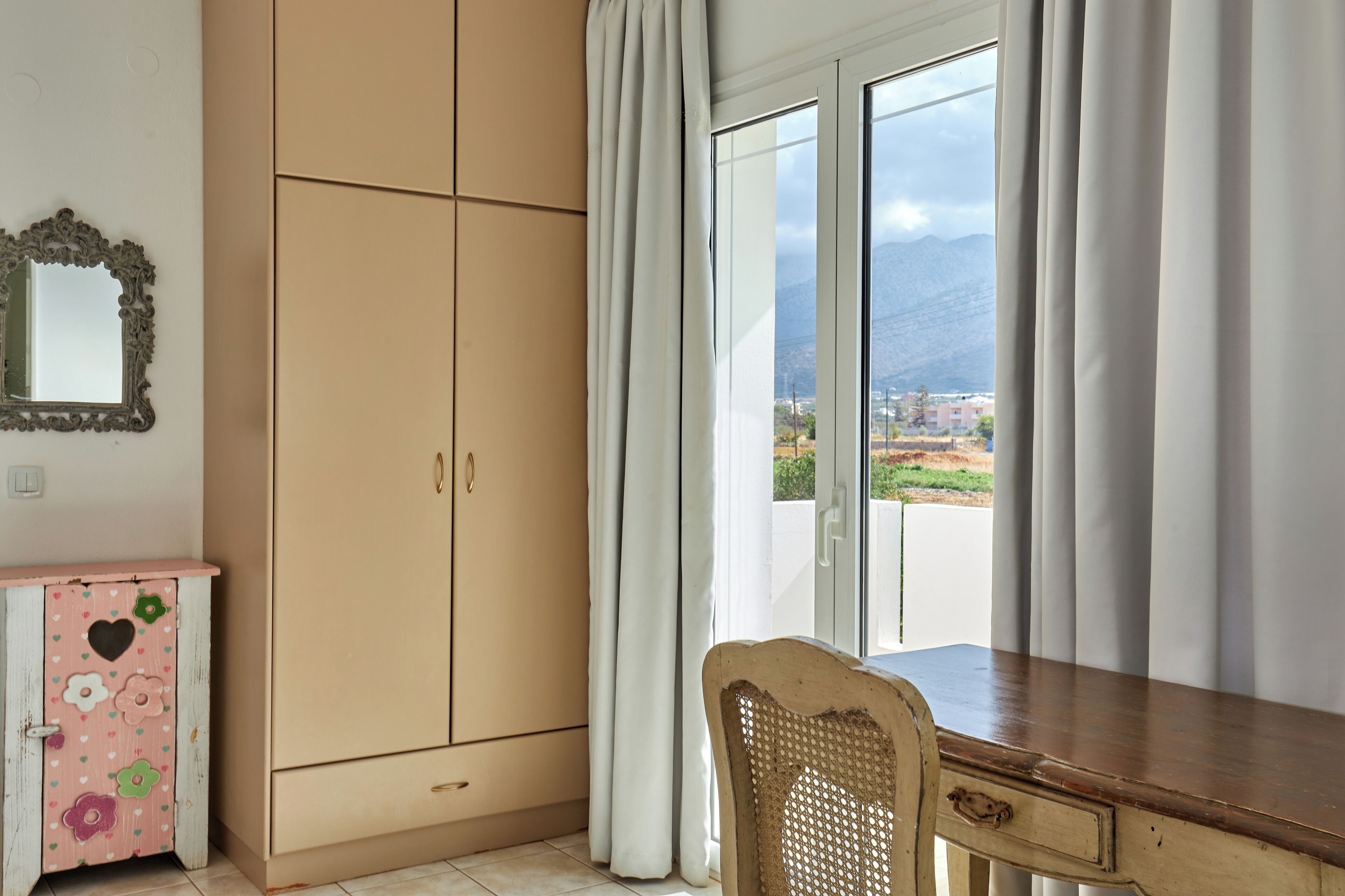 Ein Zimmer mit einem Holztisch, einem rosa Schrank mit floralem Muster und einer Glastür, die auf einen Balkon mit Bergblick führt.