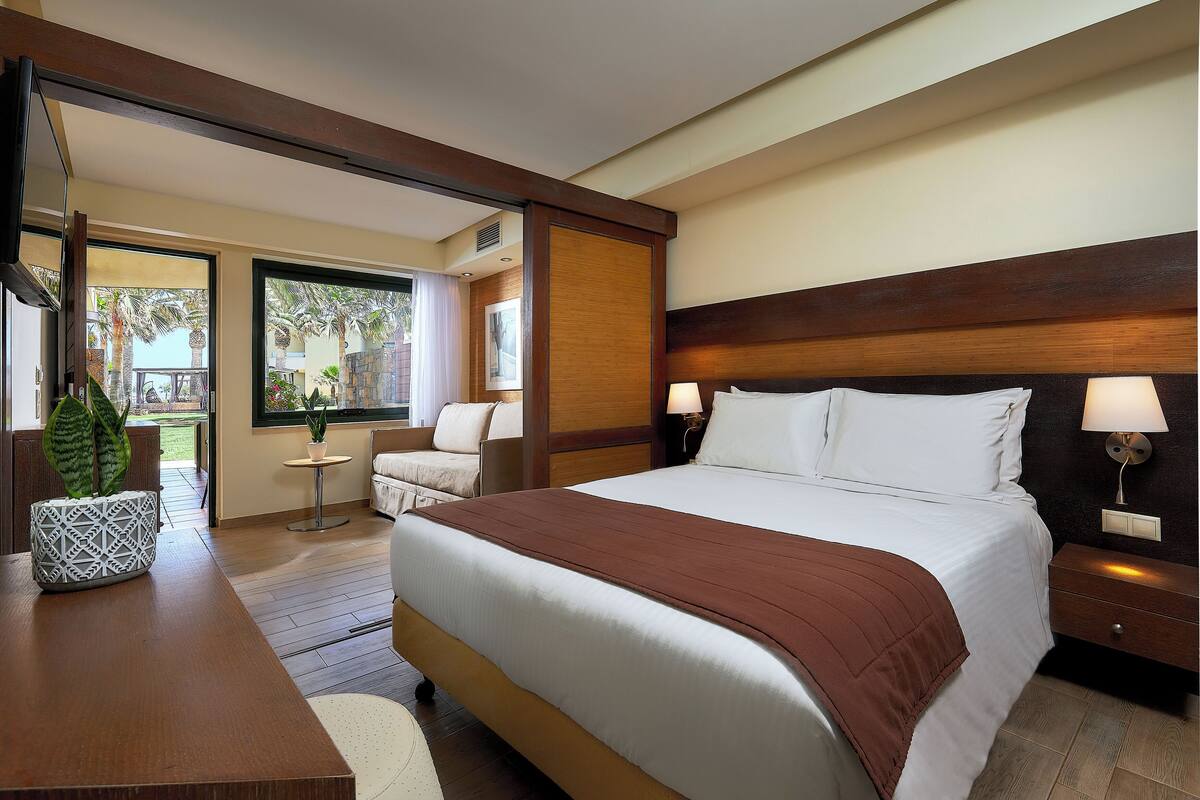 A hotel room with a large bed, a desk with a plant, a chair, a small table, and a view of the outdoors.