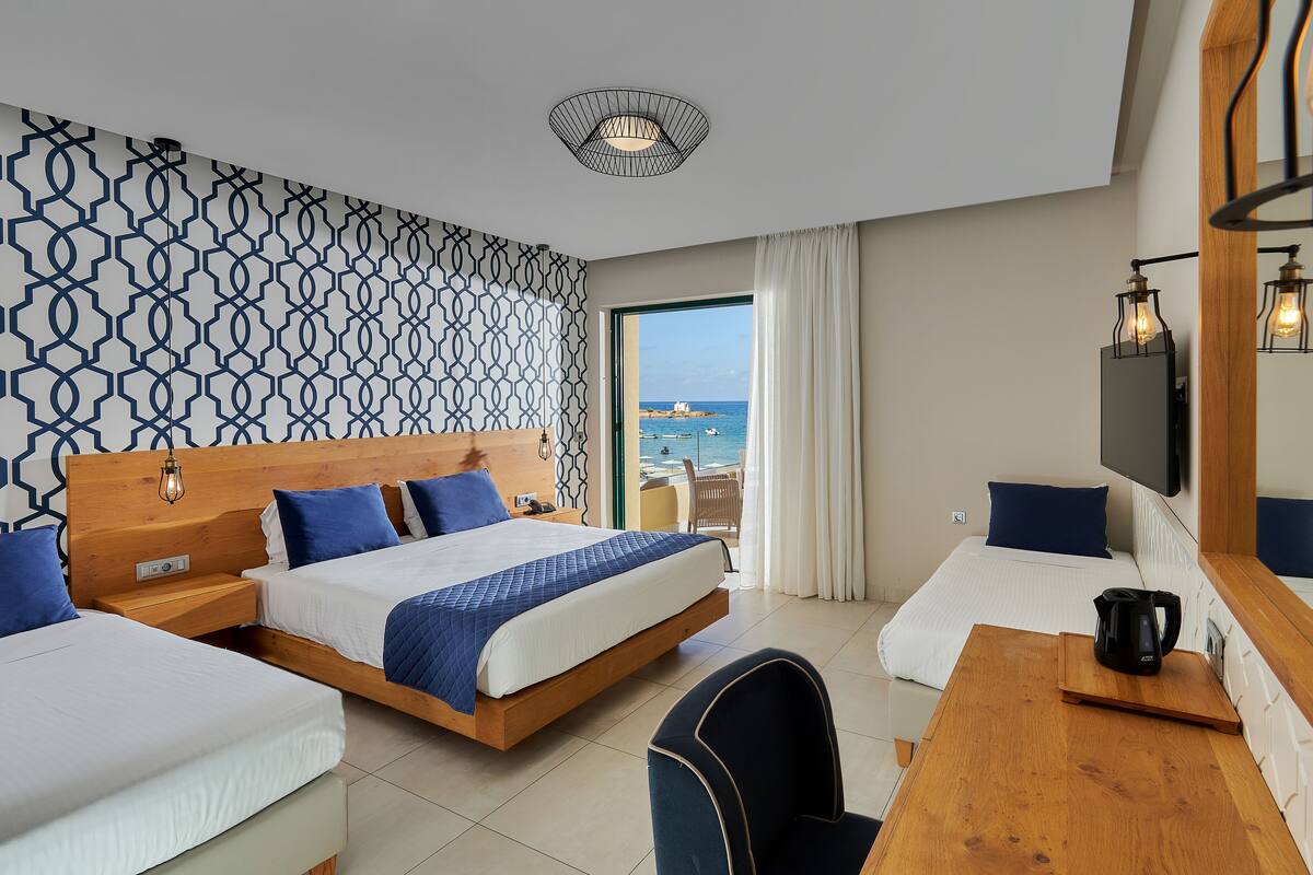 A hotel room with two beds, a desk, a chair, a TV, and a view of the sea.