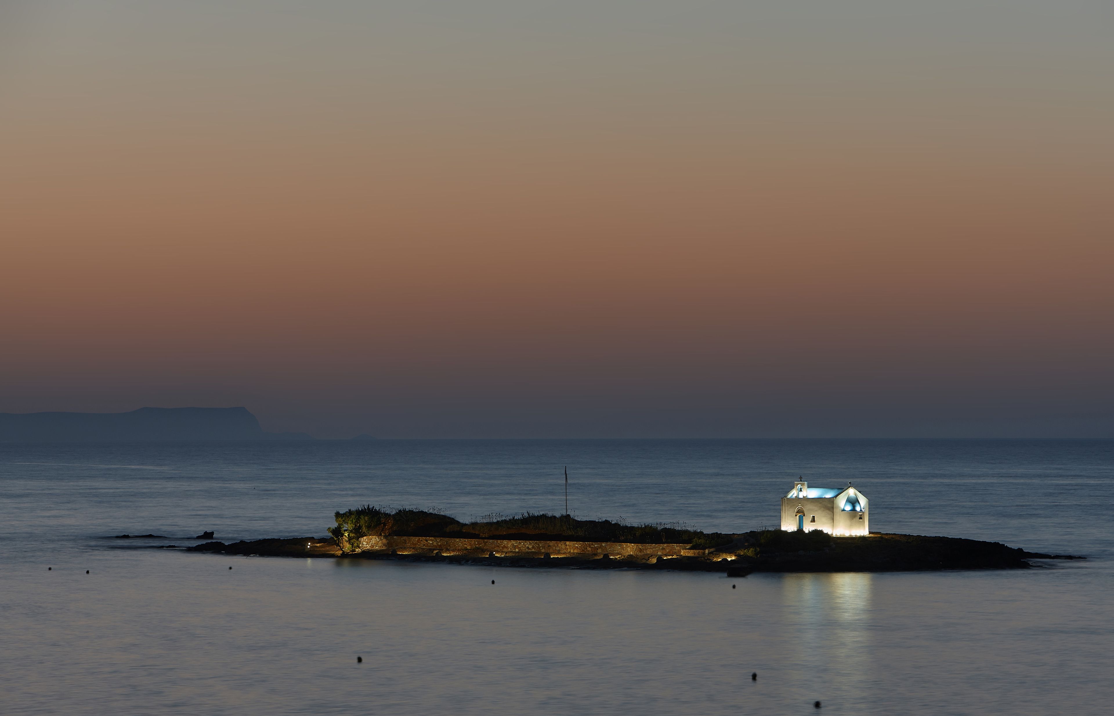 Une petite île avec un seul bâtiment, probablement un phare, illuminé sur fond de ciel crépusculaire.