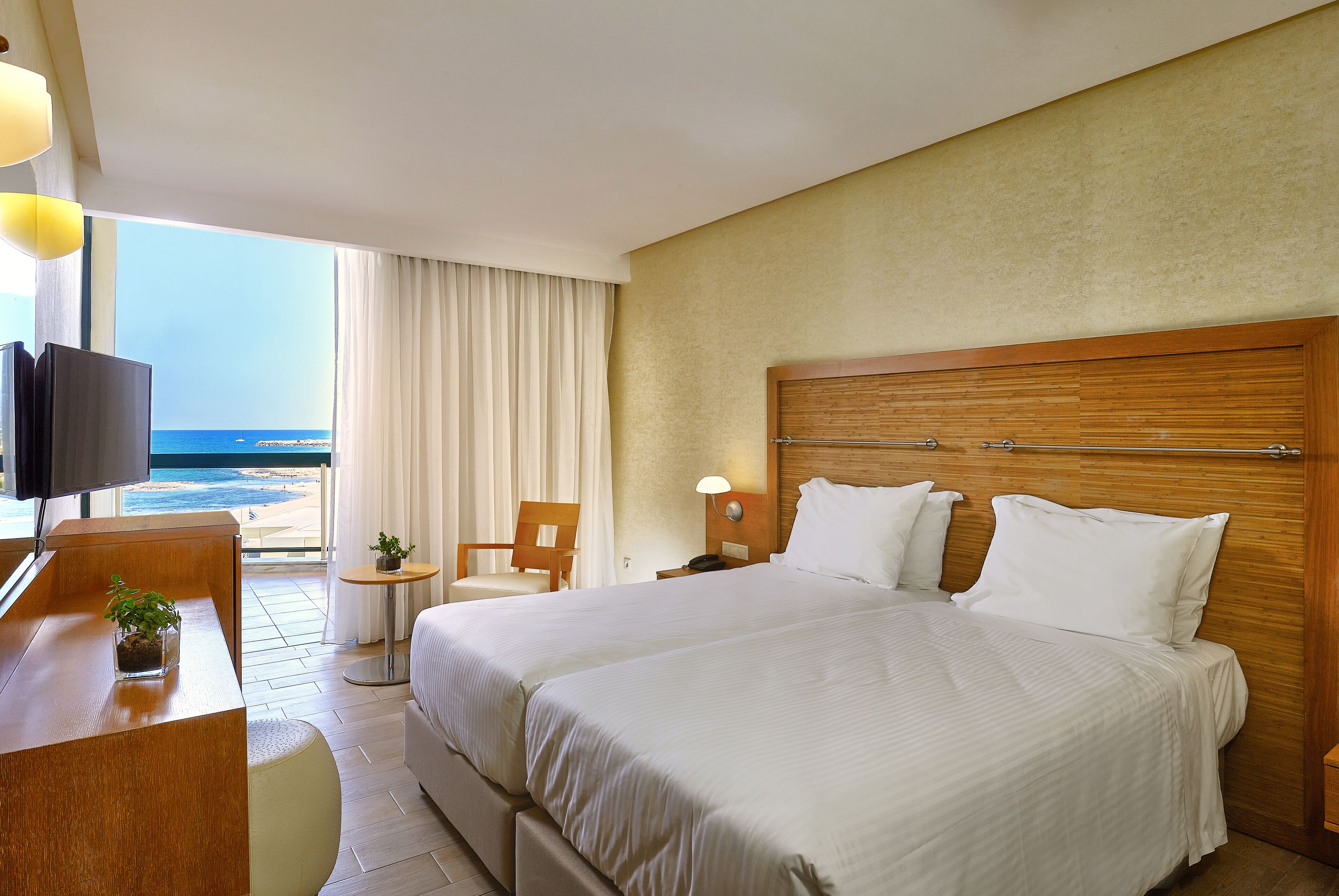 A hotel room with a large bed, a desk with a TV, a chair, a small table, and a view of the ocean.