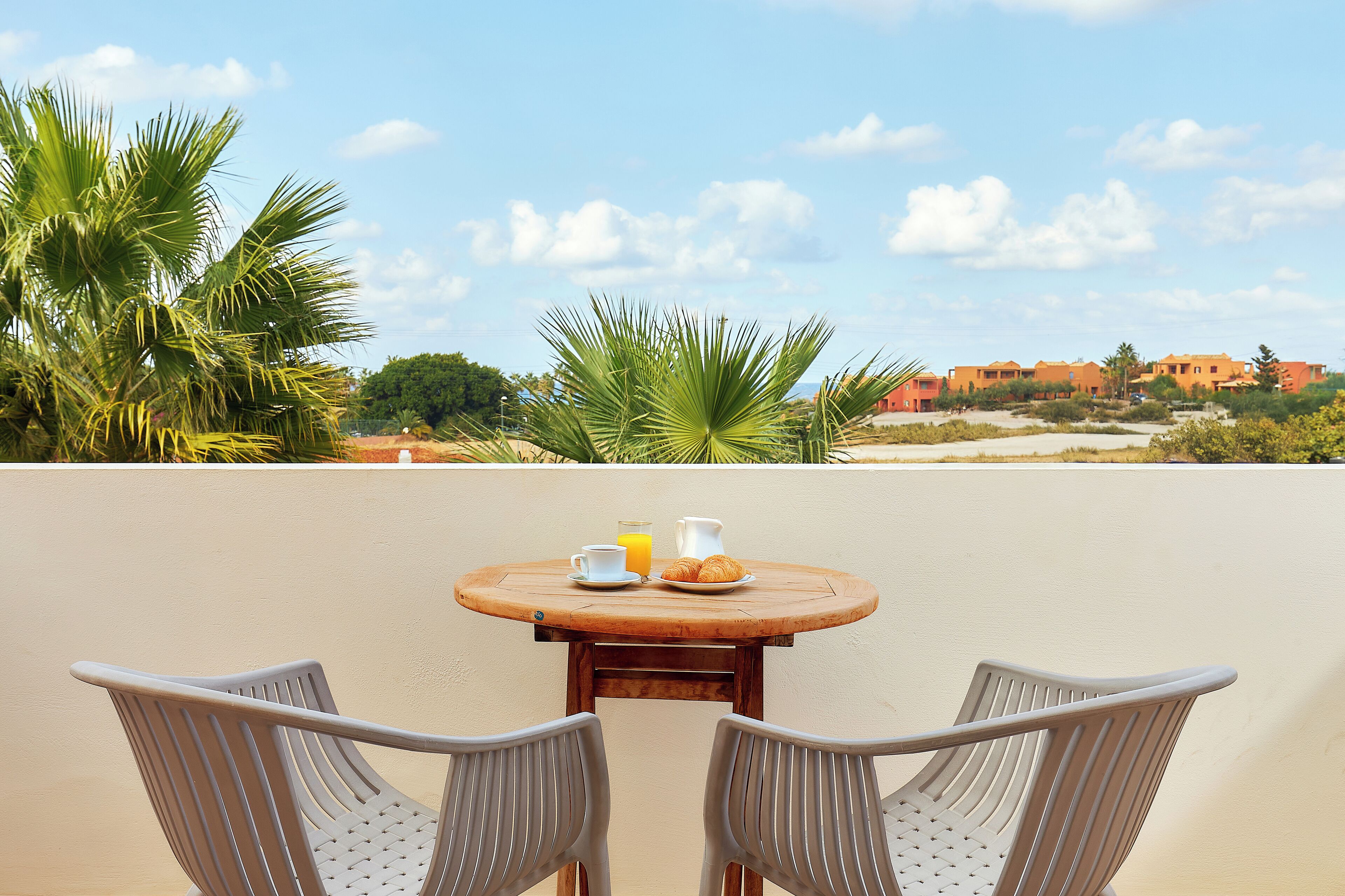 Une table ronde en bois, accompagnée de deux chaises, un verre de jus d’orange et un croissant, sur un balcon offrant une vue sur des palmiers et des immeubles.
