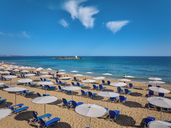 A beach with rows of blue lounge chairs and white umbrellas, a clear blue sky, and calm sea.