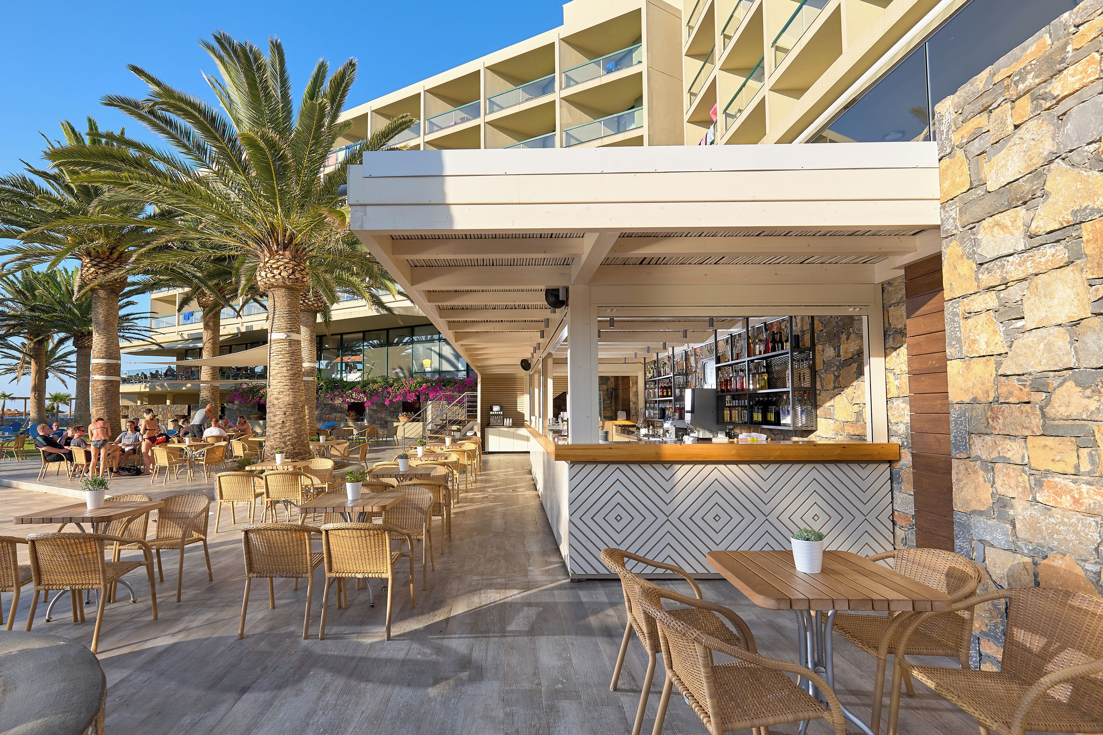 Un área de estar al aire libre con mobiliario de mimbre y palmeras, un bar con pared de piedra de fondo y un edificio moderno con balcones.