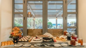 Una mesa de buffet con diversos postres y bebidas, un centro de mesa decorativo, y vistas al exterior a través de amplias ventanas.