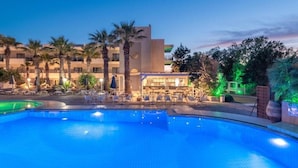 Una piscina con sillones de playa y sombrillas, un edificio de diseño moderno y palmeras.