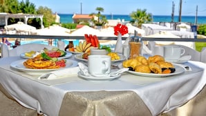 Mesa dispuesta con artículos de desayuno, como huevos, cruasanes y fruta, con vistas a una piscina y una playa.