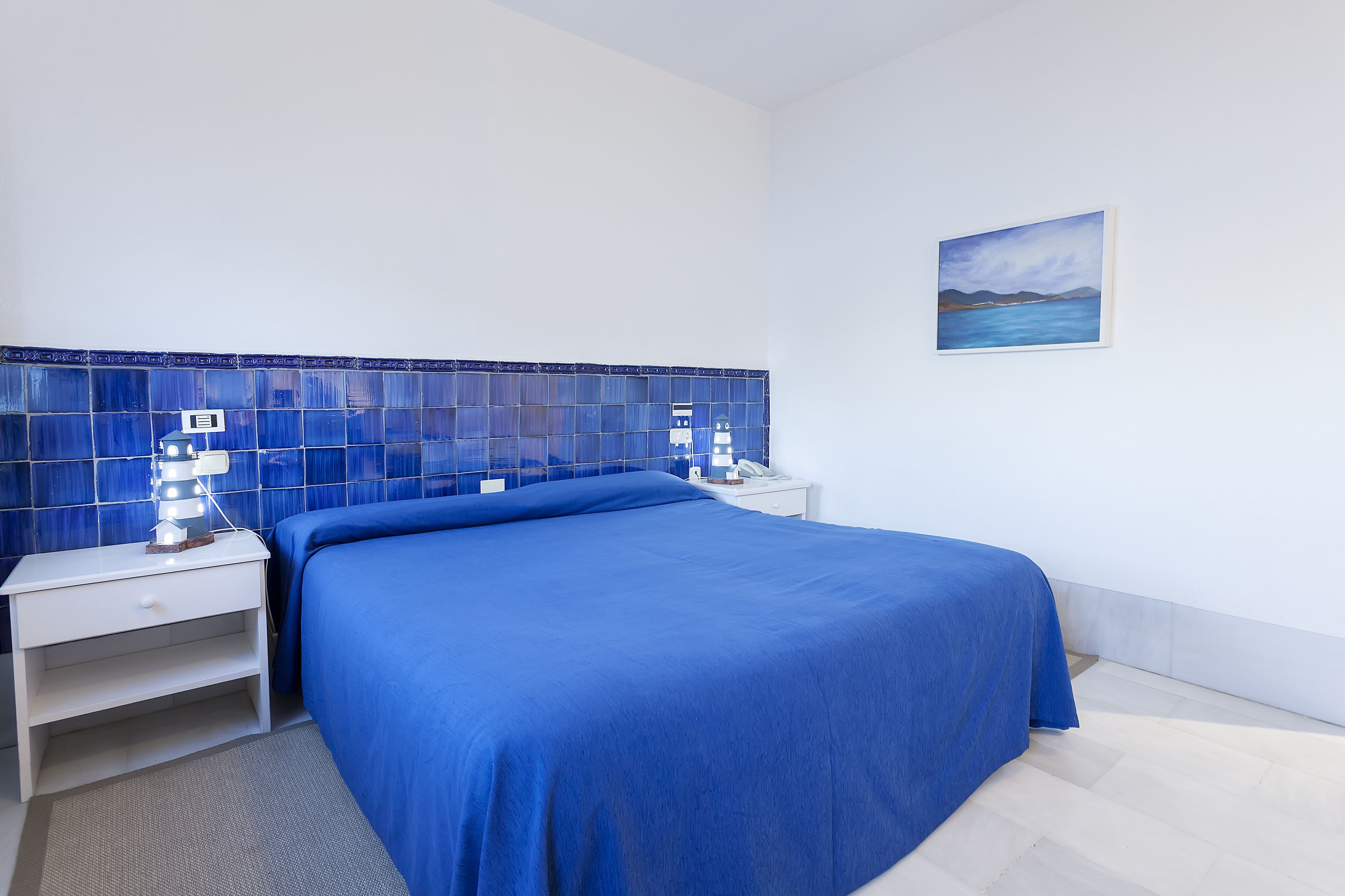 A bedroom with a bed, a nightstand, a wall-mounted lamp, and a painting.