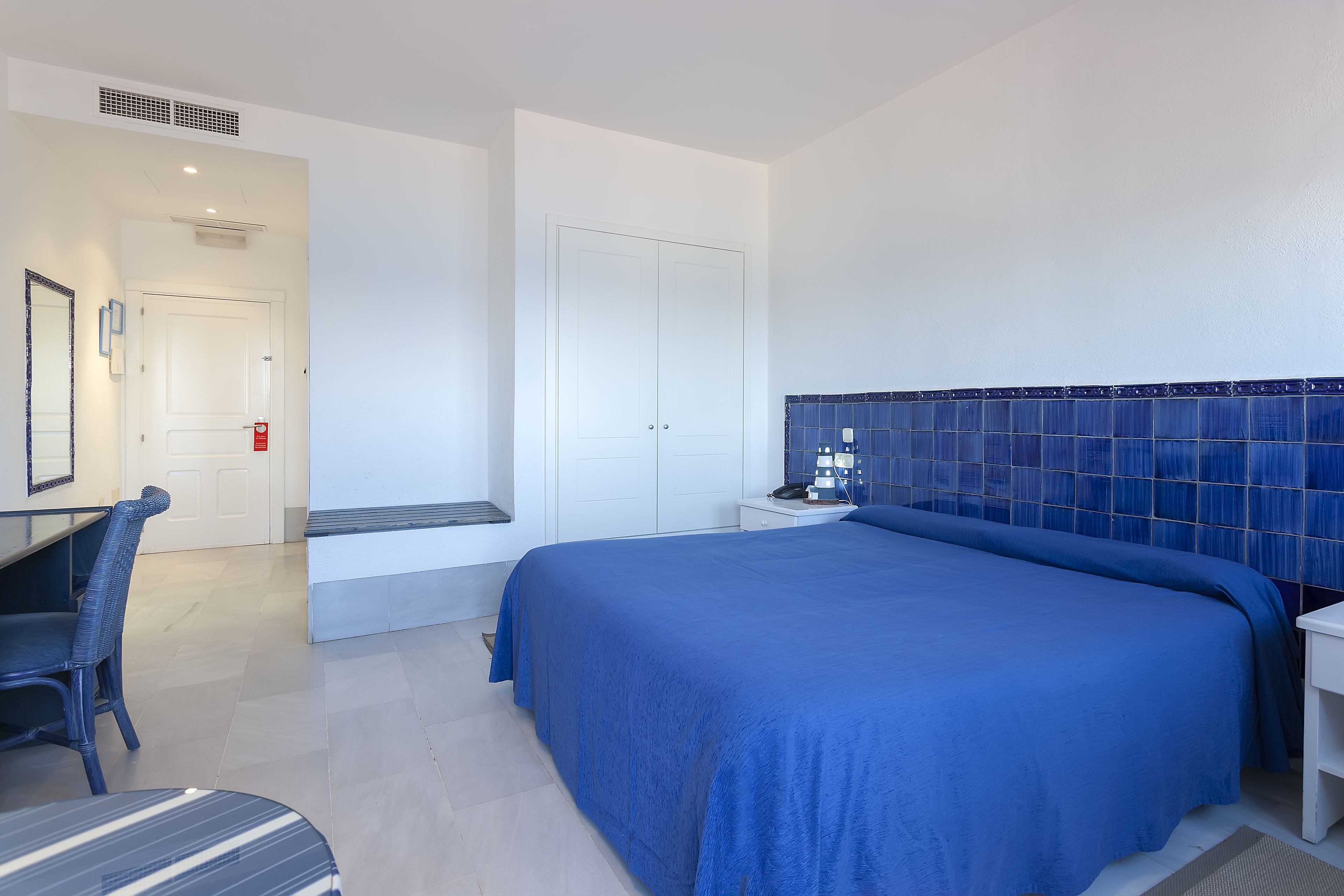 A hotel room with a large bed, a desk with a chair, a bedside table, and a tiled wall.