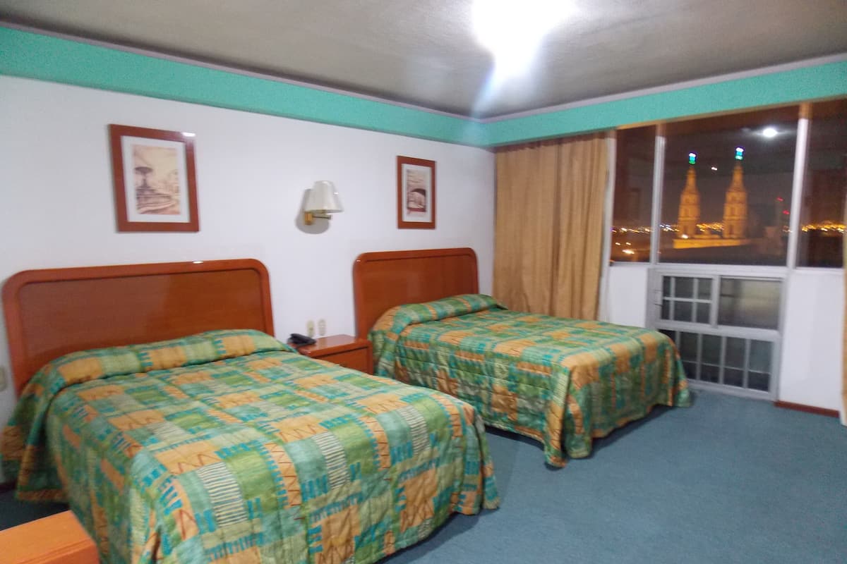 A hotel room with two beds, a nightstand, a lamp, and a window with a view of the cityscape.