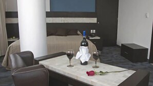 A hotel room with a bed, a bedside table with a bottle of wine and glasses, a rose, and a chair. - Hotel Real Rex (León)