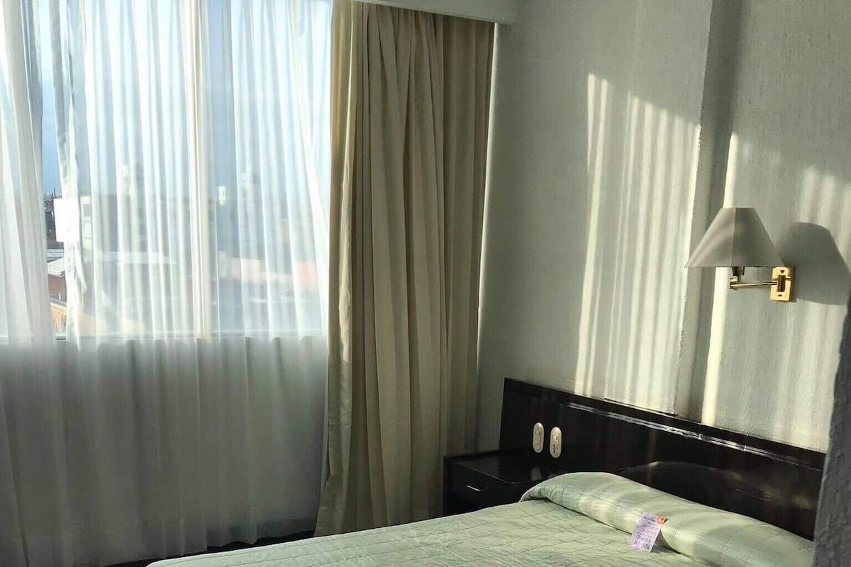 A hotel room with a bed, a nightstand, a window with curtains, and a wall-mounted light fixture.