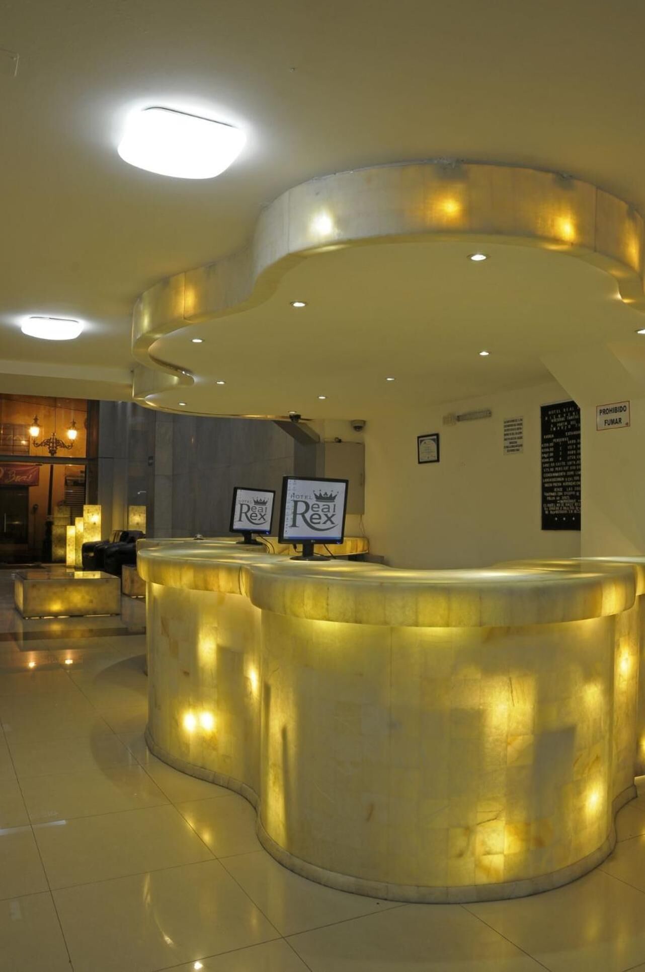 Zona de recepción de un hotel con un mostrador curvo, iluminada por luces empotradas en el techo. Hay dos monitores que muestran el nombre del hotel, y un cartel de prohibido fumar es visible en la pared.