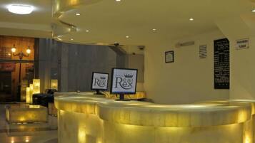 Zona de recepción de un hotel con un mostrador curvo, iluminada por luces empotradas en el techo. Hay dos monitores que muestran el nombre del hotel, y un cartel de prohibido fumar es visible en la pared.