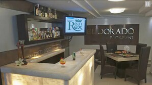 A bar area with a TV screen displaying the hotel name, a dining area with a table and chairs, and a bar stocked with various bottles. - Hotel Real Rex (León)