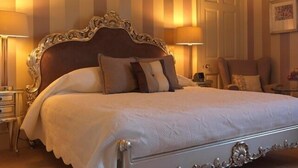 A classic four-poster bed with a white bedspread, flanked by two bedside tables with lamps, a chaise lounge, and a small table with a vase of flowers. - Ballathie Sportsmans Lodge (Perth)