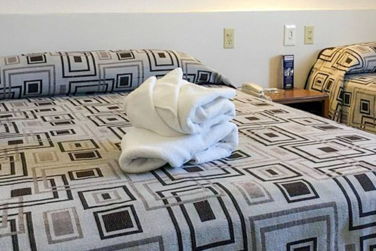 Una cama con una colcha de estampado geométrico, una toalla blanca doblada cuidadosamente y una mesita de noche con un teléfono y una caja.