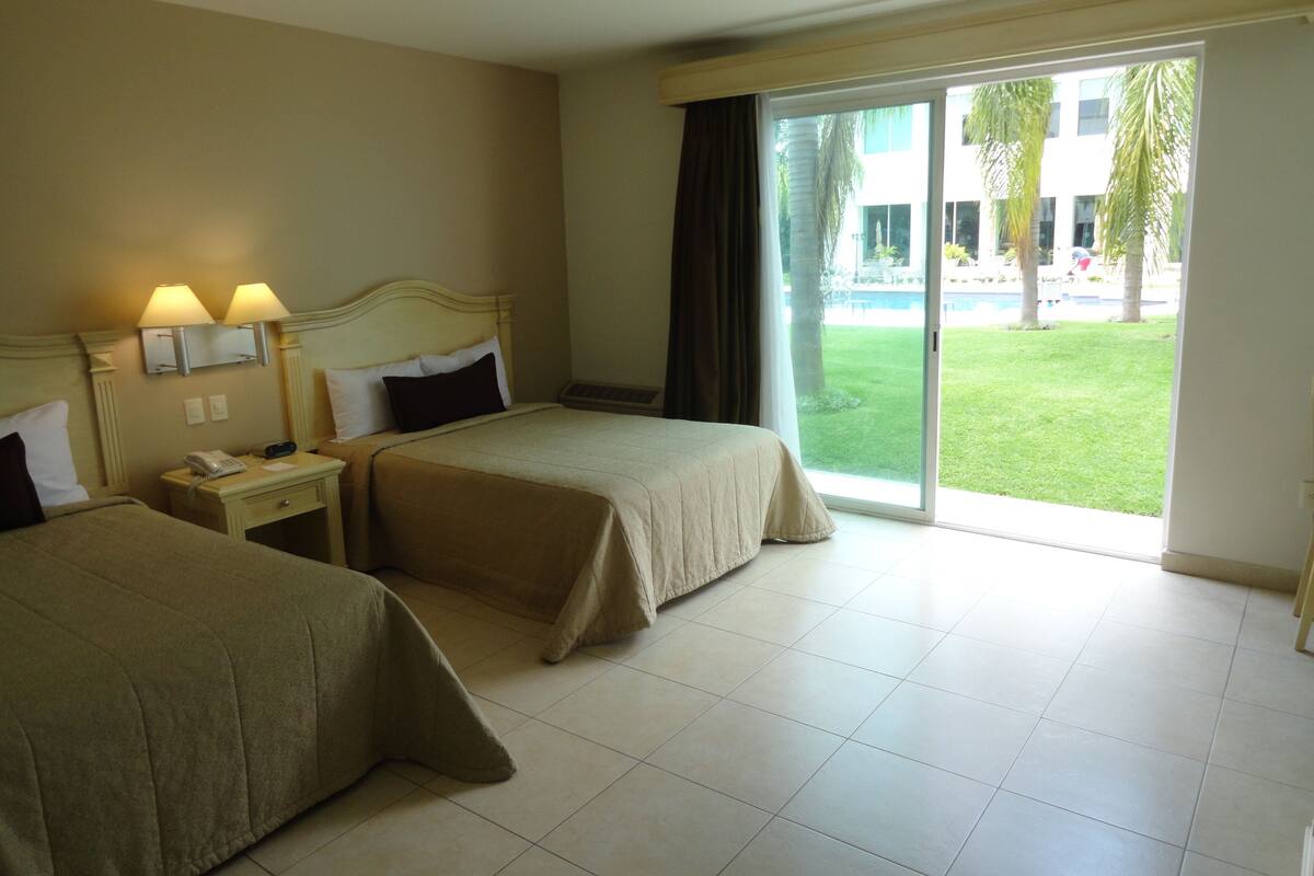 A hotel room with two beds, a nightstand, a telephone, and a window with a view of a pool and greenery.