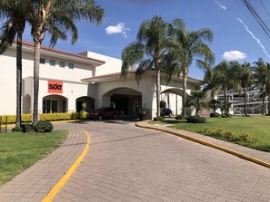 A building with a 'Sixt rent a car' sign, a covered entrance, and a parking area with cars. - Hotel Real de Minas Bajio (León)