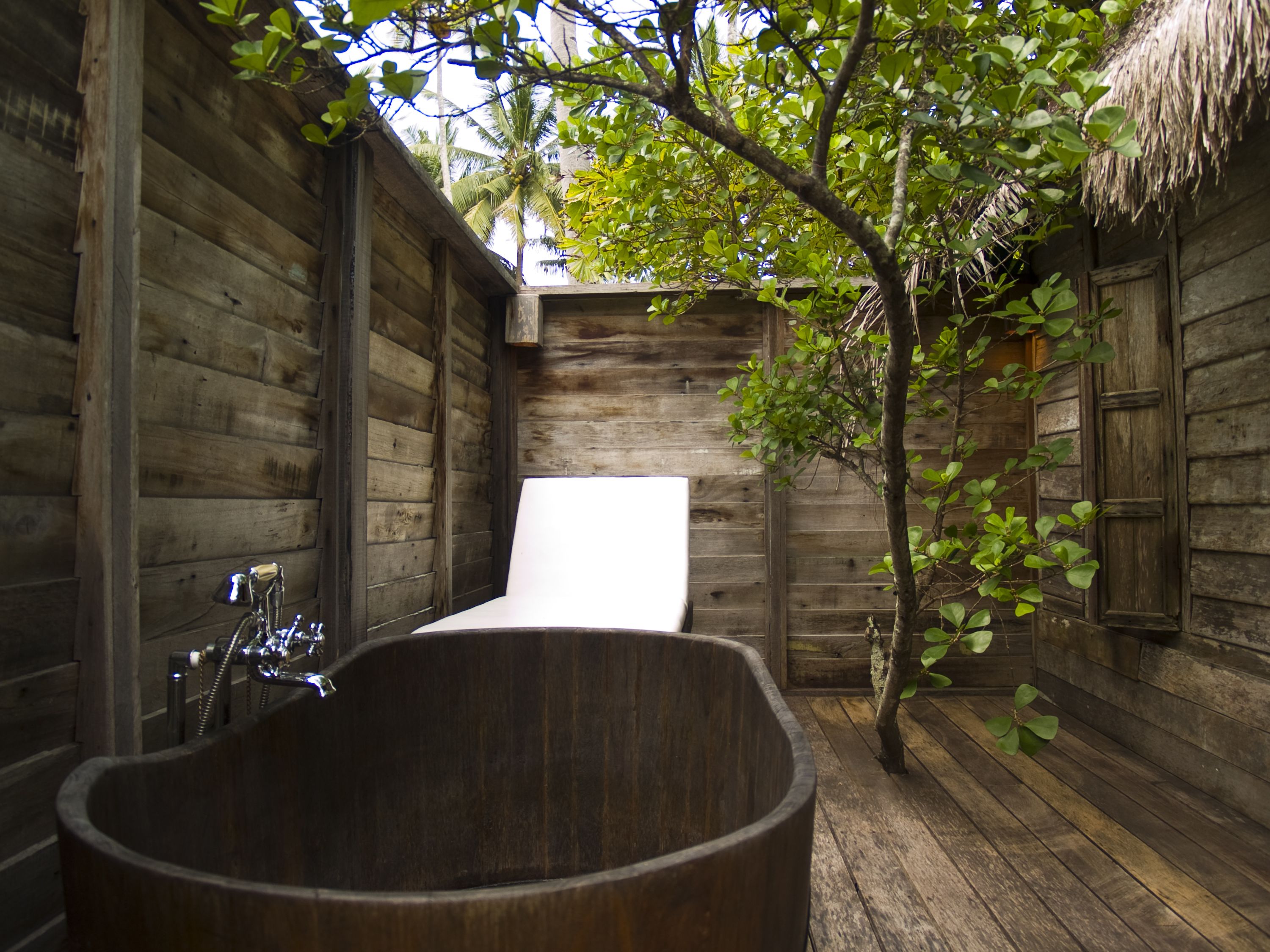 Un baño al aire libre de madera con una bañera grande, una zona de ducha y un techo de paja.