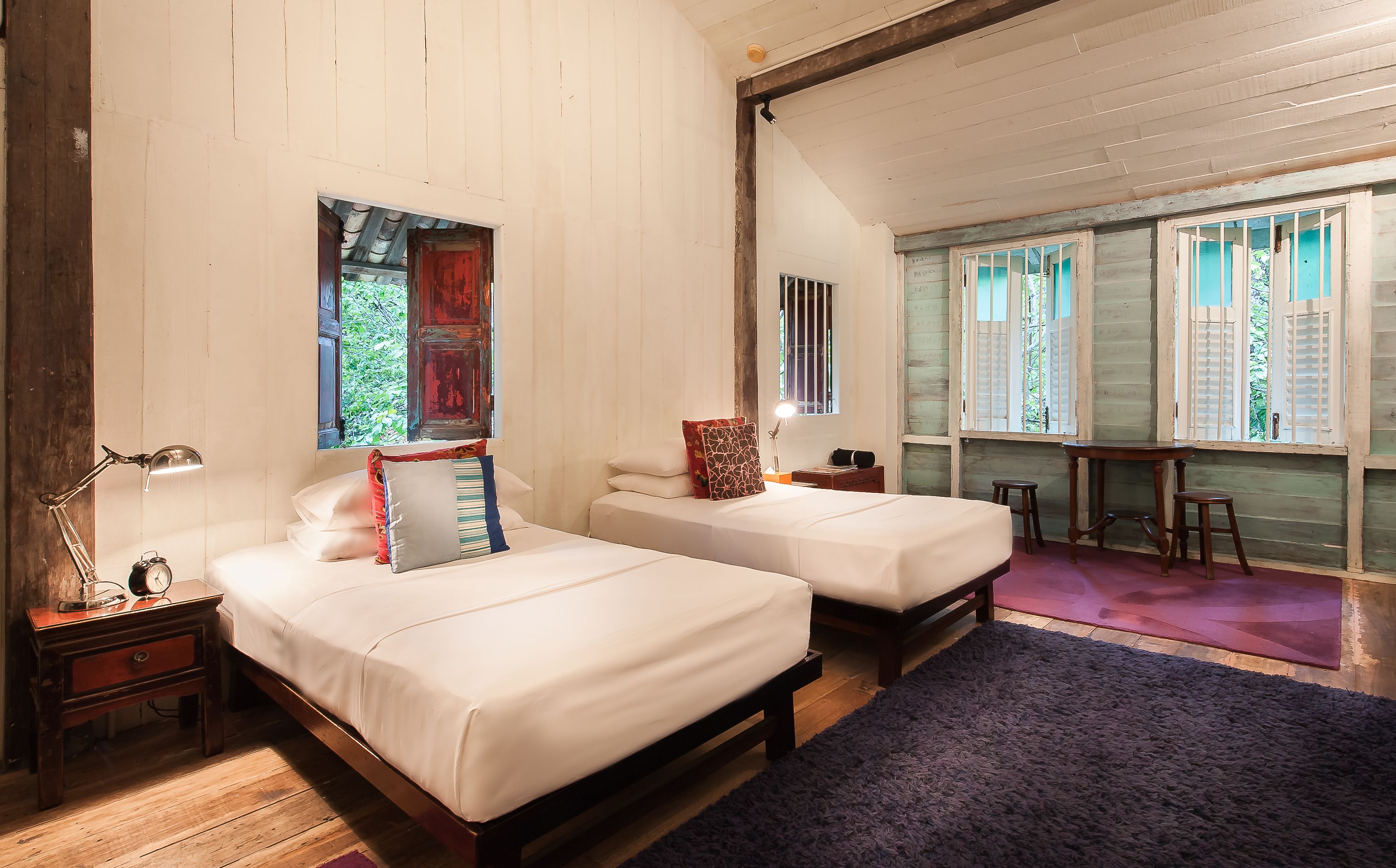 A room with two beds, a nightstand with a lamp, a small table, and a window with bars.