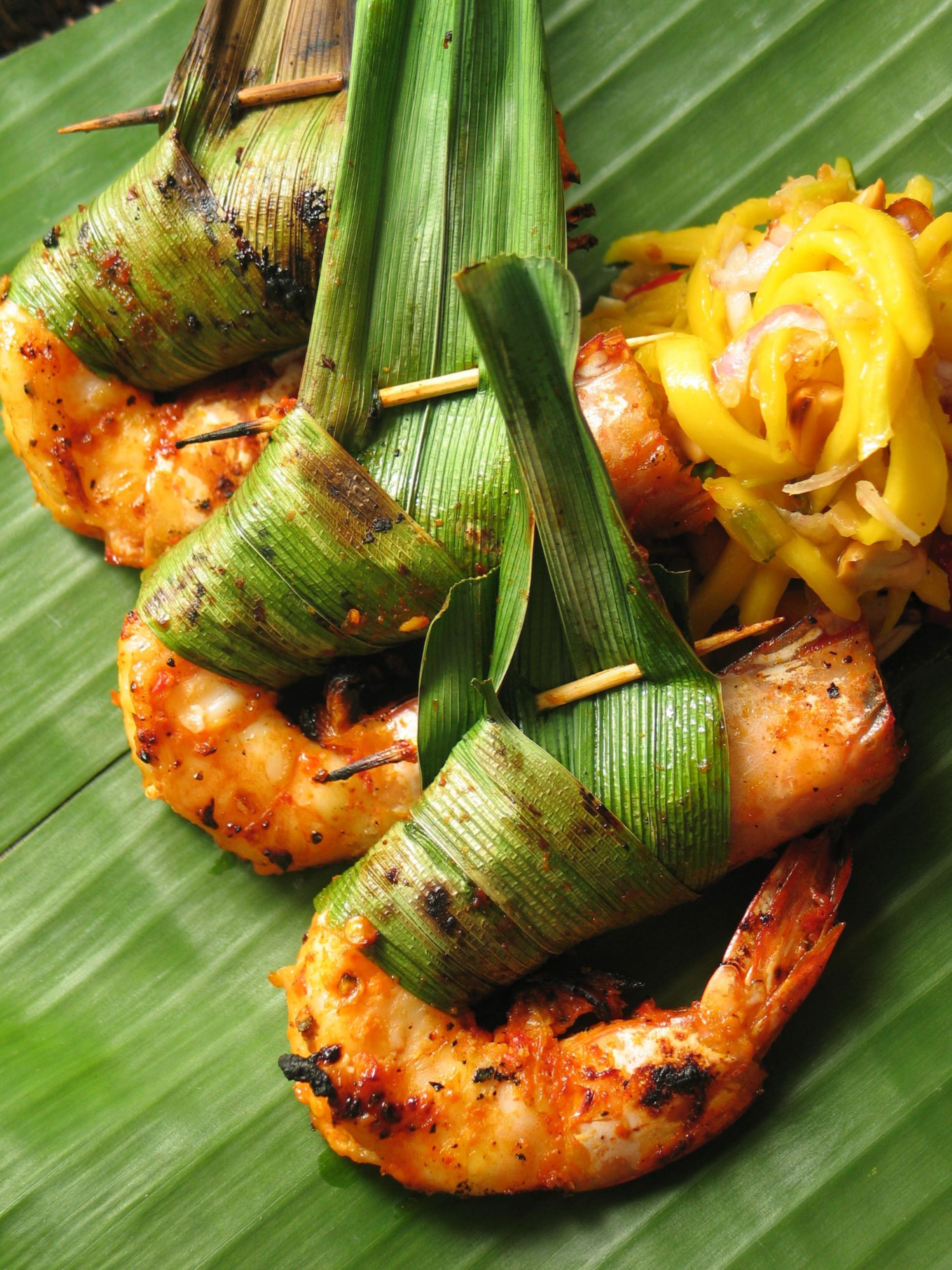 Grilled shrimp wrapped in banana leaves, served with a side of yellow vegetables.