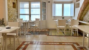 Un espace repas avec des tables et des chaises blanches, un mur en pierre et de grandes fenêtres.