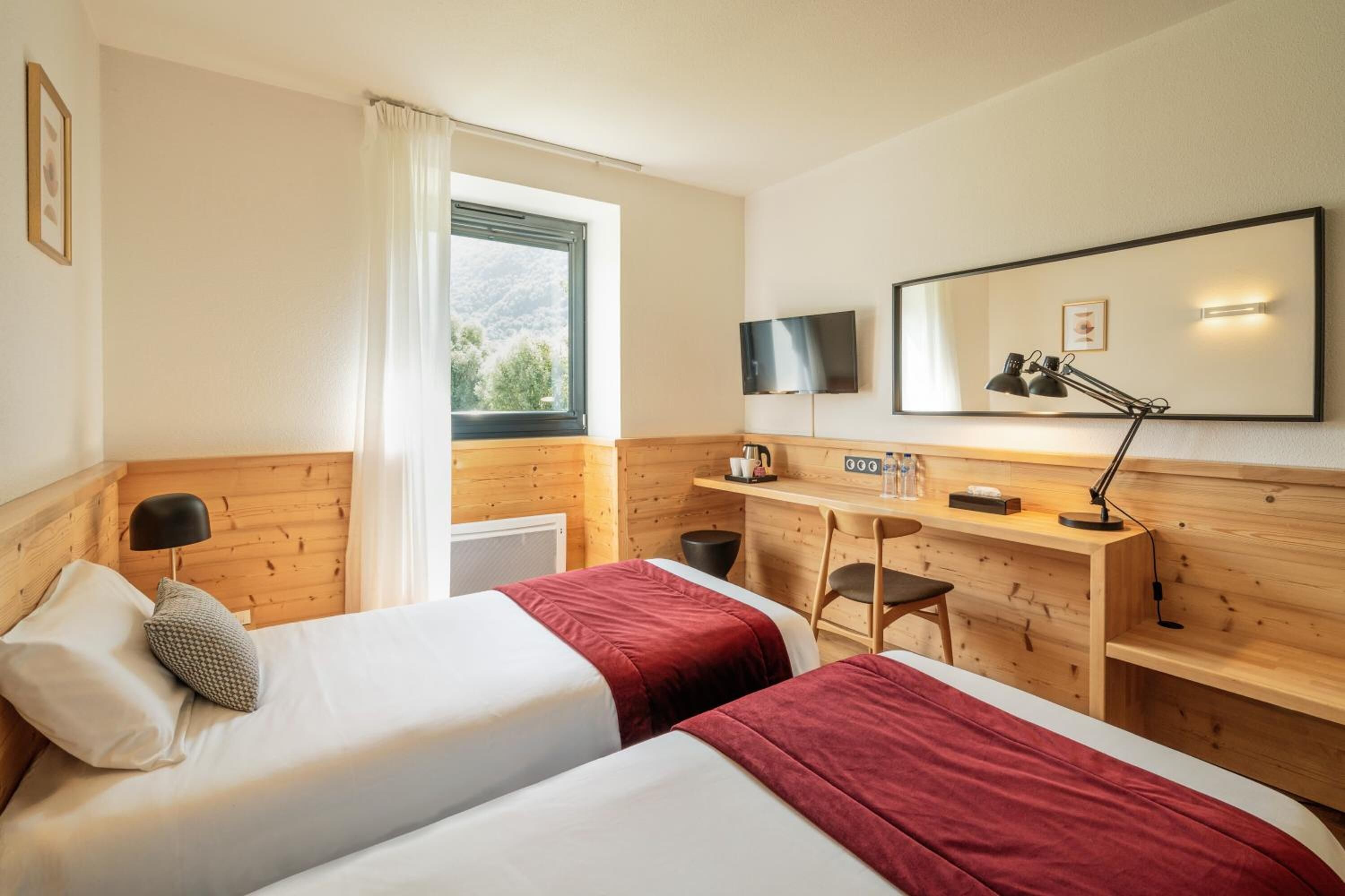 Une chambre d’hôtel avec deux lits, un bureau, une chaise, une télévision et une fenêtre donnant sur un paysage verdoyant.