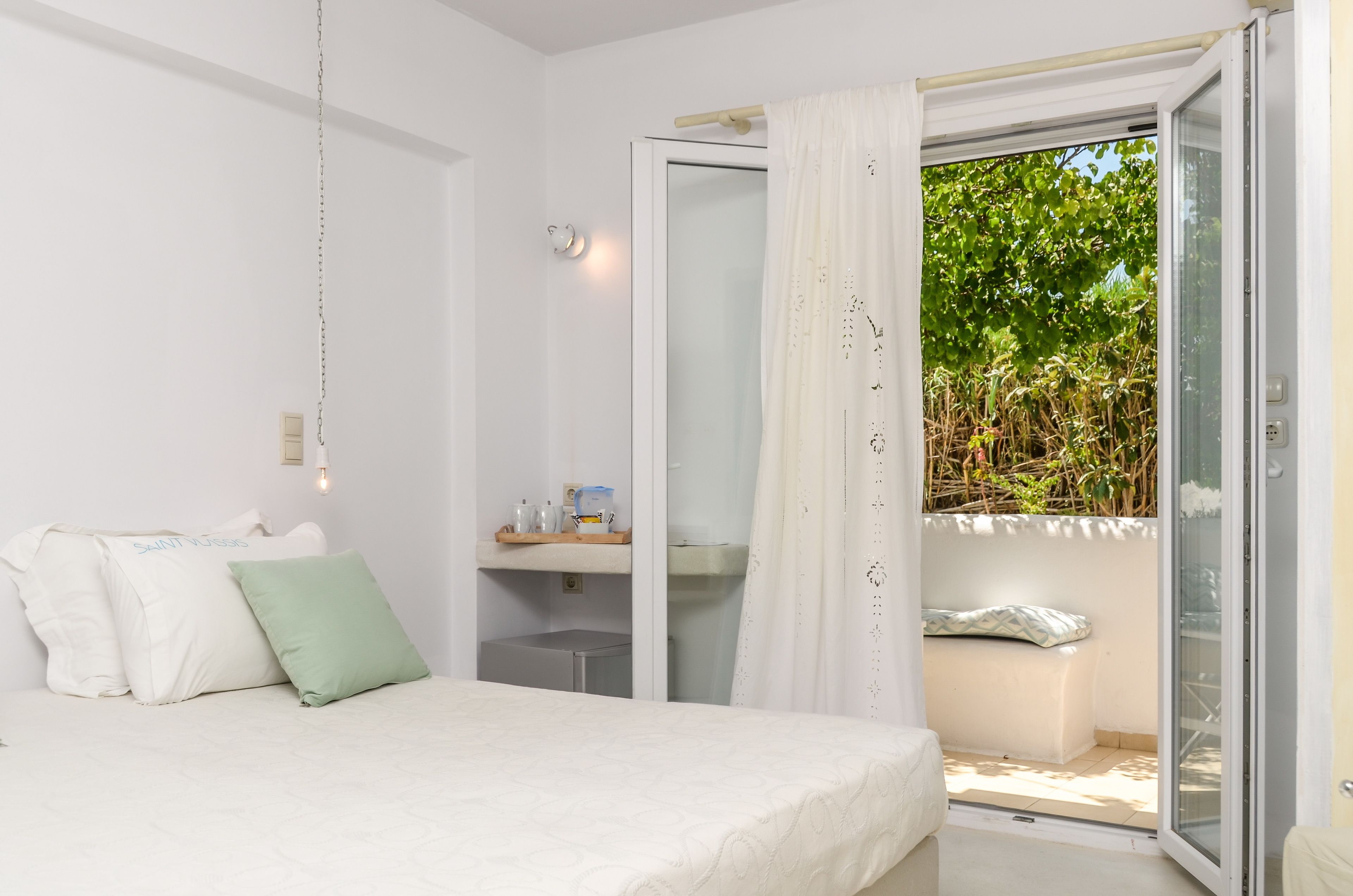 A bedroom with a bed, a nightstand, a small table with toiletries, and a sliding glass door leading to a balcony with greenery outside.