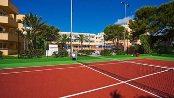 A tennis court with a net, surrounded by greenery and buildings in the background.