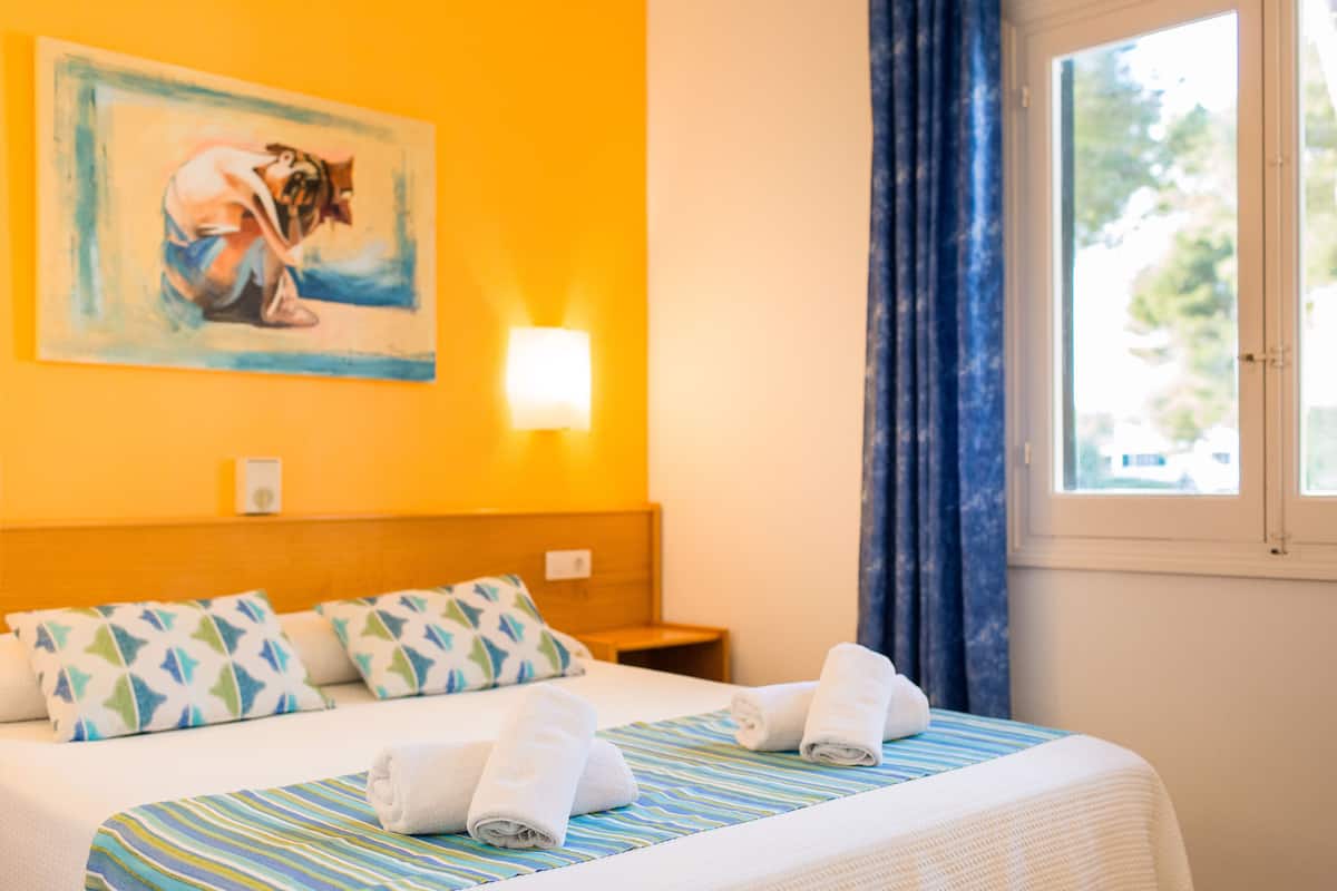 Ein Hotelzimmer mit Bett, Kissen, einem Gemälde, einem Nachttisch und einem Fenster mit Vorhängen.