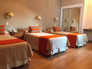 Habitación de hotel con tres camas, cada una con una colcha estampada y almohadas naranjas. Hay mesitas de noche con lámparas, un teléfono y un espejo.
