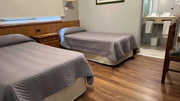 Habitación de hotel con dos camas, piso de madera, un cuadro en la pared y baño con espejo y perchero para toallas.