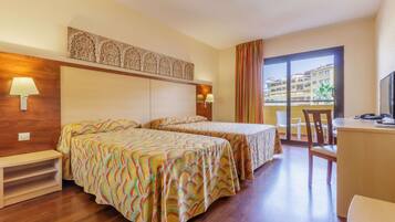 A hotel room with two beds, a desk, a chair, a TV, and a balcony with a view of buildings.