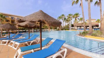 Een zwembadterras met ligbedden en rieten parasols, palmbomen en een gebouw op de achtergrond.