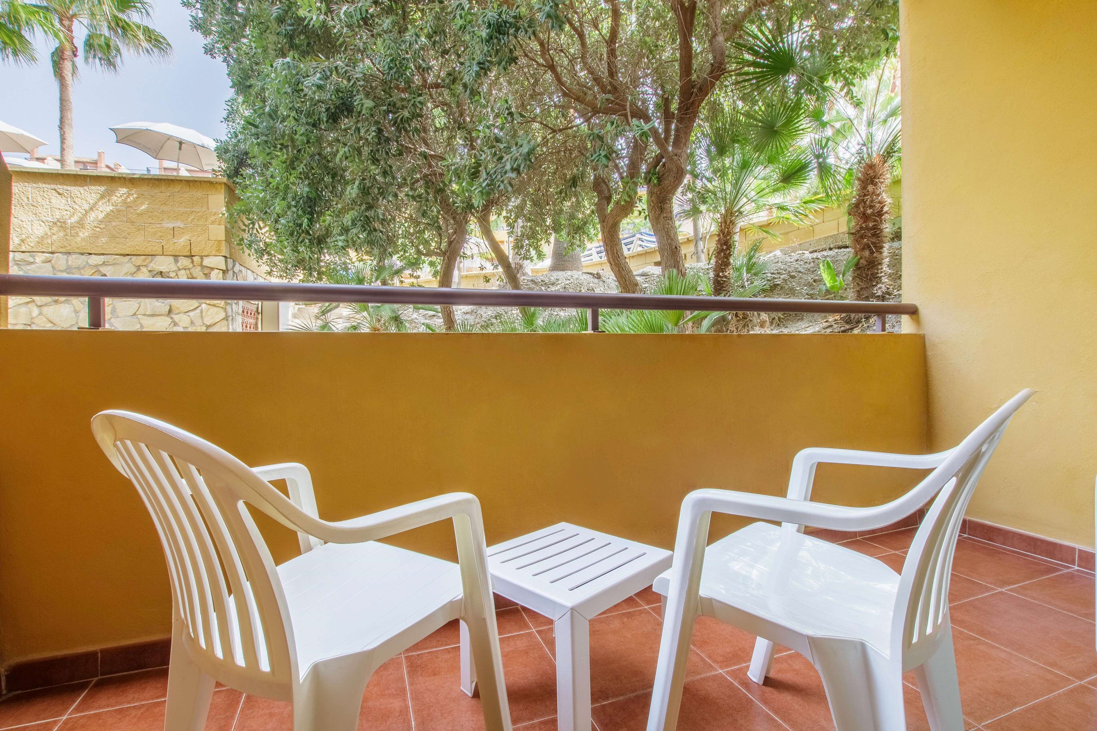 A balcony with two white plastic chairs and a small table, overlooking a garden with trees and a stone wall.