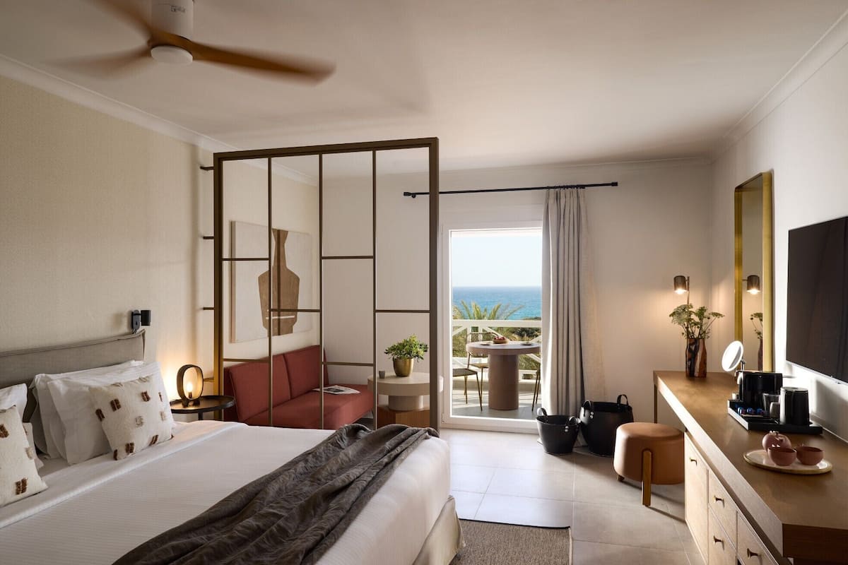Habitación de hotel con cama, escritorio, silla, televisor, balcón con vistas al mar y ventilador de techo.