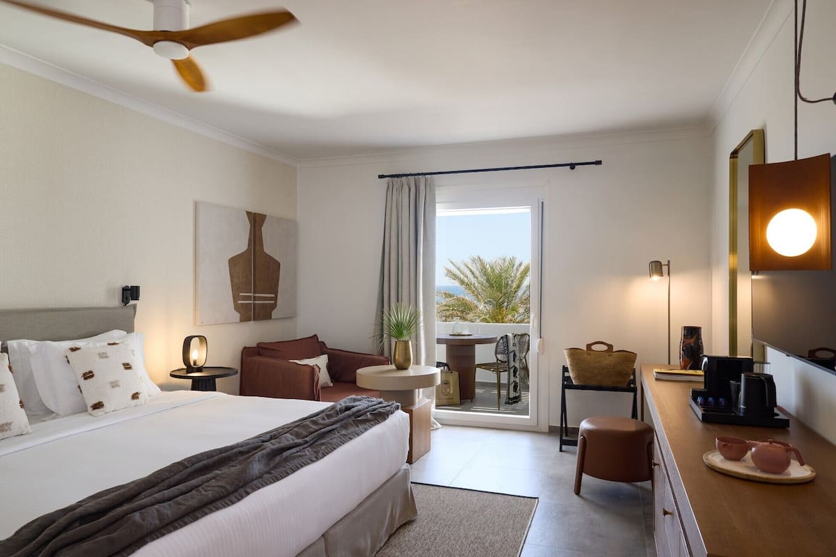 Habitación de hotel con cama, mesita de noche, lámpara y vistas al exterior.