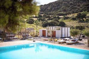 Uma área de piscina com espreguiçadeiras e um pequeno edifício, cercada por vegetação e colinas.
