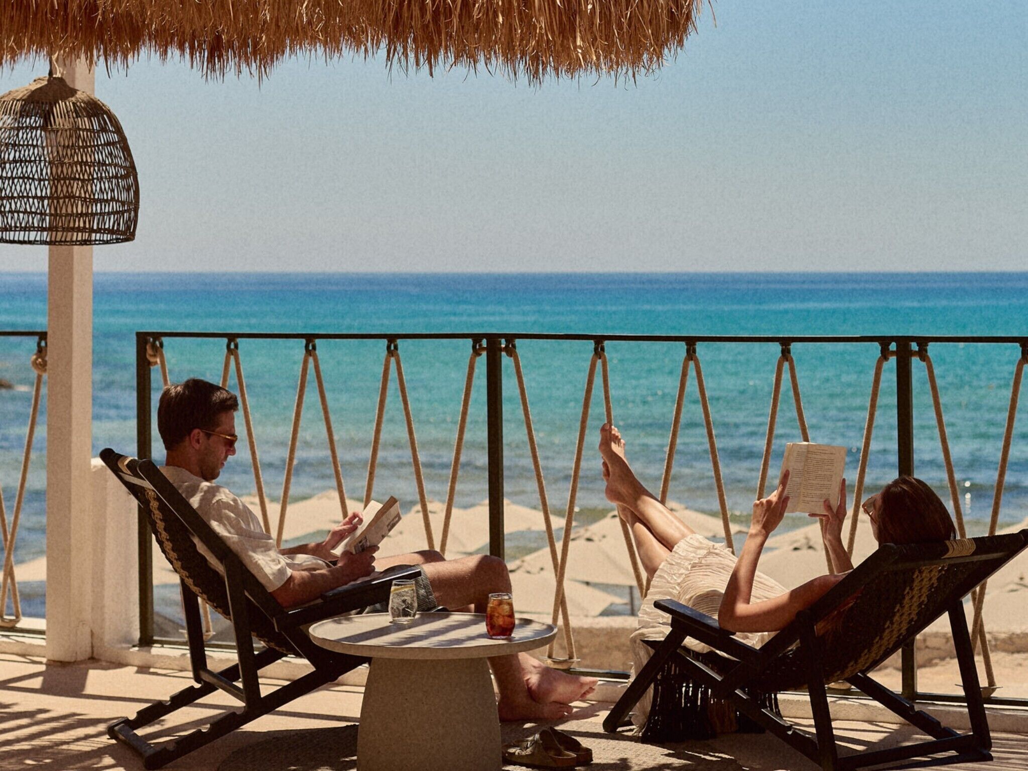 Eine Strandlage mit Schaukelstuhl, rundem Tisch und einem Reetdachschirm.