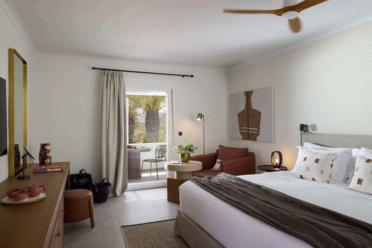 Habitación de hotel con cama, escritorio, silla, mesa, lámpara y vista a palmeras por la ventana.