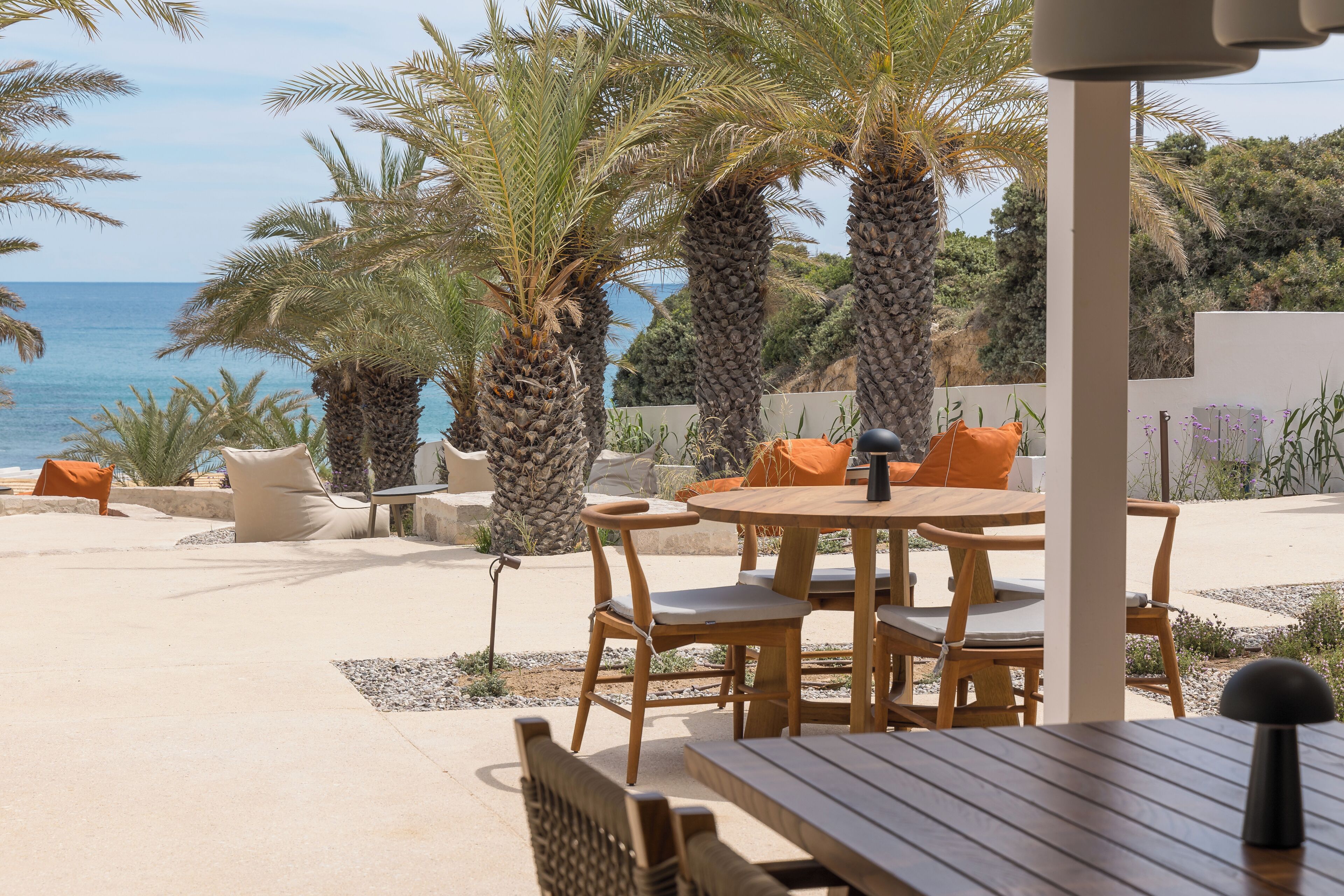 Un área de comedor exterior de madera con vistas al mar, rodeada de palmeras y una pared blanca.