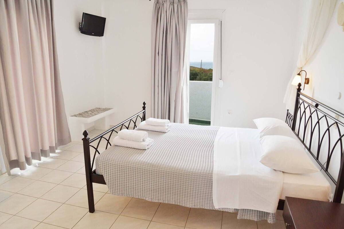 A neatly made bed with white linens, a small bedside table, a TV mounted on the wall, and a view of the sea through a door.